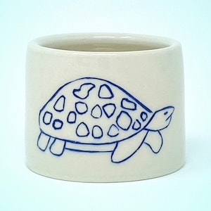 May include: A white ceramic mug with a blue outline of a turtle with a patterned shell. The turtle is facing right and has a simple design.