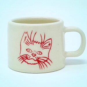 Puede incluir: Una taza de cerámica color crema con un diseño de cara de gato rojo. La cara del gato es un dibujo lineal simple con ojos expresivos, bigotes y una boca ligeramente abierta. La taza tiene un asa redondeada y un cuerpo recto.