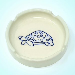 以下が含まれることがあります： クリーム色のセラミック灰皿、青いカメのイラスト付き。 灰皿は円形で、タバコを置くための3つの切り欠きがあります。 カメのデザインは、水玉模様の甲羅が特徴です。
