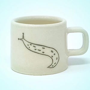 May include: Off-white ceramic mug with a simple, square handle. A gray illustration of a slug is on the side of the mug. The slug has a dotted pattern. The mug is a cylindrical shape.