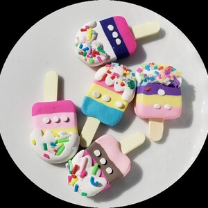 Puede incluir: Cuatro adornos de arcilla con forma de paleta de colores vivos con chispas y glaseado blanco. Cada paleta tiene un esquema de color diferente y está decorada con puntos blancos.