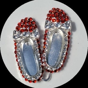 May include: Pair of silver-toned ballet slipper brooches, encrusted with red and clear rhinestones. Each slipper features a bow and a mirrored interior, creating a sparkling, elegant accessory. The brooches are set against a white background.