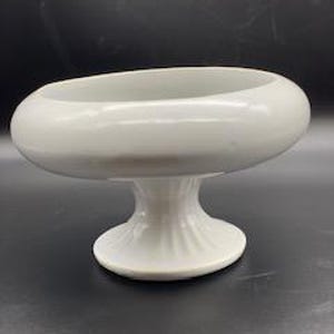 May include: White ceramic pedestal bowl with a rounded, oval shape. The bowl has a smooth, glossy finish and a raised base.