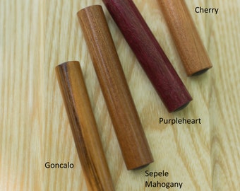 Könnte beinhalten: Fünf Holzstiftchen aus verschiedenen Holzarten, darunter Goncalo, Sepele Mahagoni, Purpleheart und Kirsche. Die Stiftchen sind diagonal auf einer hellen Holzoberfläche angeordnet.