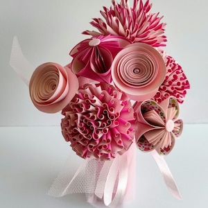 Può includere: Un bouquet di fiori di carta rosa in un vaso rosa con un fiocco di tulle bianco. I fiori sono fatti di carta e hanno una varietà di forme e dimensioni.