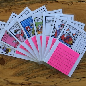 May include: A collection of humorous cartoon cards with various illustrations and text, each encased in a clear plastic sleeve. Bright pink sticky notes are attached to some of the cards. The cards are arranged on a wooden surface.