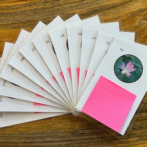 May include: A set of twelve white cards with black numbers and a pink sticky note on the top card. The top card has a pink flower with green leaves in a circle.