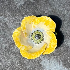 May include: A ceramic flower sculpture with bright yellow petals and a detailed centre. The flower has a textured surface and is set against a grey background. The sculpture is a decorative art piece.