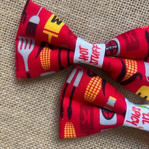 May include: Red bow tie with a white label that says "Hot Buttaff" and a black label that says "Black" with a red and yellow corn on the cob, a spatula, and a grill brush pattern.