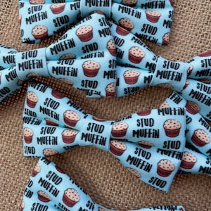 May include: Several light blue bow ties with a repeating pattern of muffins and the words "STUD MUFFIN" in black. The bow ties are arranged on a textured, tan surface.