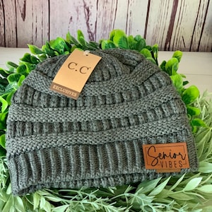 May include: A dark green knit beanie with a brown leather patch that says "Senior Vibes" and a brown tag that says "C.C. Exclusives".