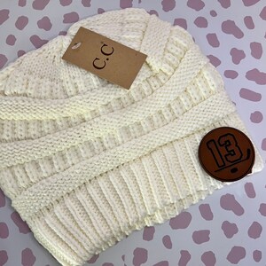 May include: A white knitted beanie with a brown leather patch that says "13" on it. The beanie has a cable knit pattern and is folded over at the top.