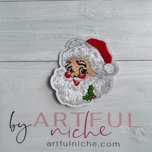 May include: Embroidered Santa Claus patch with a red hat, white beard, and rosy cheeks. The patch includes a small sprig of holly and is set against a light wood grain background. The text "by ARTFUL niche" and "artfulniche.com" are also visible.