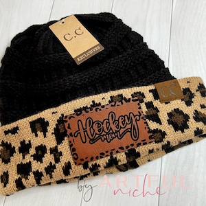 May include: Black and tan knit beanie with a brown leather patch that says "Hockey Mom". The beanie has a leopard print pattern on the bottom half.