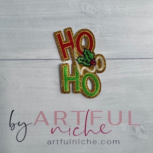 May include: A glittery Christmas decoration with the words "HO HO" in red and green, outlined in gold. A sprig of holly with red berries is between the words. The text "by Artful Niche" and the website address are at the bottom.