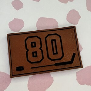 May include: Brown leather patch with black stitching and the number 80 in black outlined letters. A black hockey stick and puck are also on the patch.