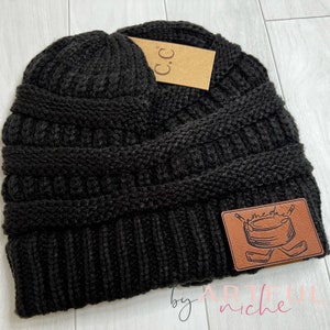 May include: Black knit beanie with a brown leather patch that says "me-day" with a hockey stick and puck design. The beanie has a fold-over cuff and is made of a soft, cozy material.