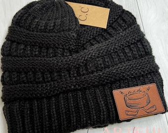 May include: Black knitted beanie with a brown leather patch that says "me-day" with a hockey stick and puck design. The beanie has a fold-over cuff and is made of a soft, cosy material.