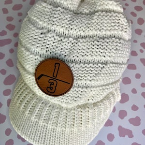 May include: White knitted beanie with a brown leather patch featuring a hockey stick and the number "13".