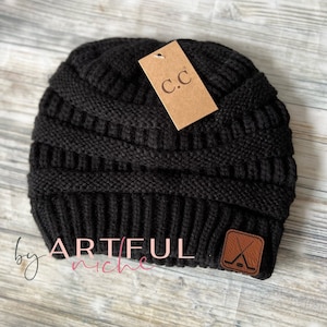 May include: Black knit beanie with a brown leather patch featuring a hockey stick and puck. The beanie has a fold-over cuff and a brown tag with the text "C.C." on it. The beanie is made by "ARTIFUL niche".