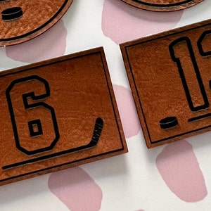 May include: Two brown leather hockey jersey patches with black numbers and a hockey stick and puck design. The numbers on the patches are "6" and "13".