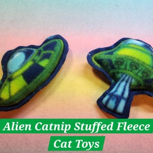 May include: Two catnip-stuffed fleece toys shaped like a UFO and a mushroom. The UFO is blue and green with a light blue window. The mushroom is green and blue with a light blue stem. The toys are on a rainbow-colored background. Alien Catnip Stuffed Fleece Cat Toys