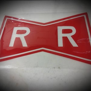 May include: A red and white emblem featuring two white "R" letters. The emblem has a bow-tie shape with a white border. The background is a glossy red color. The emblem is likely a logo or a symbol.