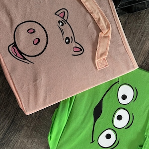 May include: Two fabric storage bins. One is light pink with a cartoon pig face design in black. The other is bright green with a cartoon alien face design in black. Both bins have matching pink handles.