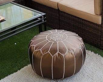 May include: A sectional sofa with beige cushions and dark brown wicker frame. A brown leather ottoman with white stitching sits on a white rug with black geometric patterns. A glass-top coffee table and decorative items complete the outdoor living space.