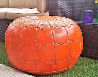 May include: An orange leather pouf with intricate stitching and a decorative top. The round ottoman is placed on a green artificial lawn, near a white rug and outdoor furniture. The pouf is a stylish accent piece.