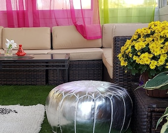 May include: Outdoor living space with a beige sectional sofa, a glass-top coffee table, and a silver ottoman. A white rug with black geometric patterns sits on the artificial grass. Yellow flowers and green plants add color.