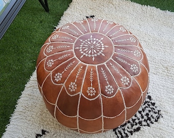 May include: A brown leather pouf with white stitching sits on a white rug with black diamond patterns. A glass-topped coffee table and a beige sectional sofa are in the background. The scene is set on a green surface, suggesting an outdoor patio.
