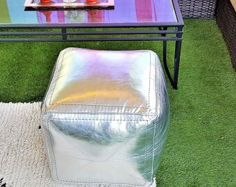May include: A silver cube-shaped ottoman with stitched seams sits on a white rug with black geometric designs. A glass-top coffee table with a black metal frame and a wicker sofa are in the background. The scene is set on artificial green grass.