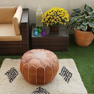 May include: An outdoor living space featuring a brown wicker sectional sofa with tan cushions, a glass-topped coffee table, and a round brown leather ottoman. A cream rug with black geometric designs sits on green turf.