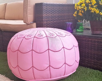 May include: A round, pink leather ottoman with a decorative stitched pattern. The ottoman is placed on a green surface, with a beige sectional sofa and a dark brown wicker storage chest in the background. A rug and plants are also visible.