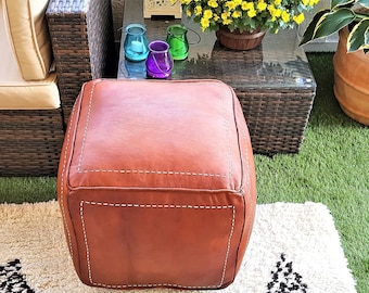 May include: A square, brown leather ottoman with white stitching details. The ottoman is placed on a white rug with a black and white geometric pattern. The background includes outdoor furniture and potted plants.