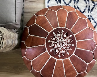May include: Round brown leather pouf with a geometric design. The pouf is made of various shades of brown leather panels stitched together with white thread. A white embroidered starburst pattern adorns the center. The pouf is a decorative home furnishing.