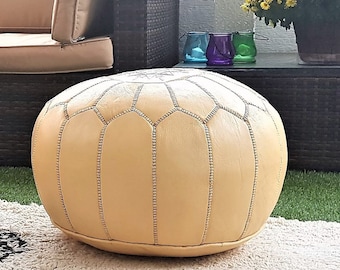 May include: An outdoor living space featuring a beige sectional sofa, a glass-top coffee table, and a round, light yellow leather ottoman. A cream rug with black geometric patterns sits on the green turf. Potted plants and flowers add color.