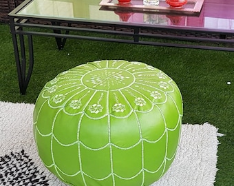 May include: A bright green round ottoman with white stitching sits on a white rug with black diamond patterns. A black metal coffee table and outdoor seating are in the background. The scene is set on artificial turf.