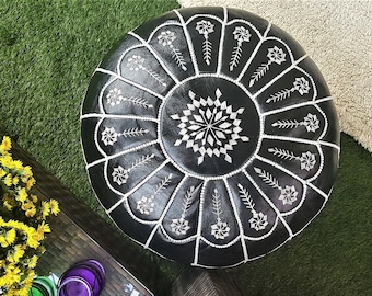 May include: A round black leather pouf with white stitching and floral patterns. The pouf is a decorative piece of furniture, perfect for a living room or bedroom. The pouf is approximately 20 inches in diameter.