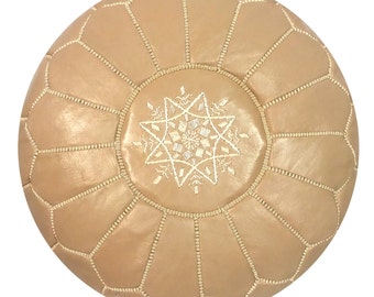 May include: A round, tan leather pouf with a geometric pattern. The pouf features a central starburst design embroidered in white thread. The edges are stitched with a lighter tan thread, creating a decorative border. This Moroccan-style pouf is a decorative home furnishing.