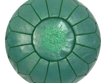 May include: A round, emerald green leather pouf with a geometric pattern. The pouf features a central embroidered design in a lighter shade of green. The stitched seams and the embroidered starburst design add texture and detail to the decorative home furnishing.