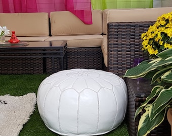 May include: A white leather pouf with a geometric design, placed on a green surface. The round ottoman is a decorative piece of furniture. The background includes a beige sofa, a glass table, and plants.
