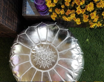 May include: A silver leather pouf with a geometric pattern and white embroidered starburst design. The round ottoman is on a green surface, with yellow flowers and a wicker table in the background. A decorative accent for any living space.