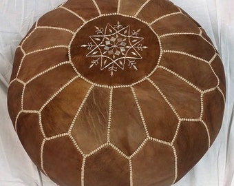 May include: Round brown leather ottoman with a geometric pattern and white stitching. The top features a white embroidered starburst design. This decorative pouf is a stylish addition to any living space.