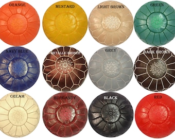 May include: A collection of round, handcrafted leather ottomans in various colors, including orange, mustard, light brown, green, navy blue, metallic bronze, grey, dark brown, cream, burgundy, black, and red. Each ottoman features a central starburst design.