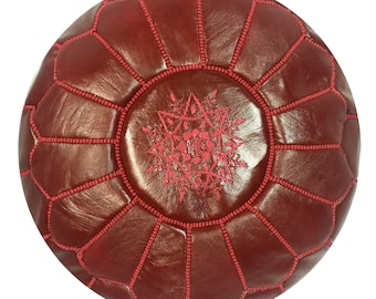 May include: Round, burgundy leather pouf with a geometric pattern. The pouf features a central embroidered starburst design in a lighter shade of red, and is stitched with red thread. This decorative ottoman is a stylish accent for any living space.