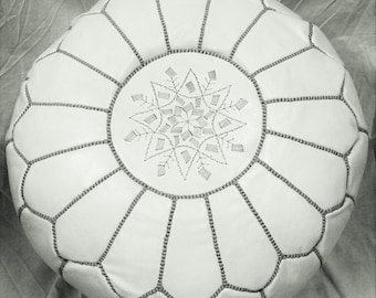May include: A round, white leather pouf with a geometric design. The pouf features a central embroidered snowflake pattern and is stitched with gray thread. The overall design is a neutral color palette, suitable for various interior styles.