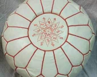 May include: Cream-colored round Moroccan pouf with a geometric pattern. The pouf features a starburst design in the center and is stitched with red thread. The pouf is a decorative and functional piece of furniture.