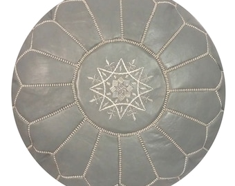 May include: A round, gray leather pouf with a starburst design embroidered in white thread at the center. The pouf is segmented into panels, each outlined with white stitching. This decorative ottoman is a stylish addition to any living space.